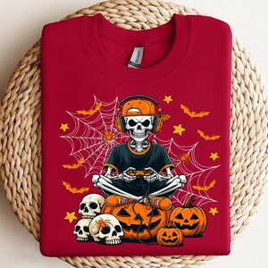 May include: A red sweatshirt with a Halloween design. The graphic depicts a skeleton wearing headphones and holding a game controller, positioned in front of pumpkins, bats, spiders, and a spiderweb. The design uses orange, black, and white.