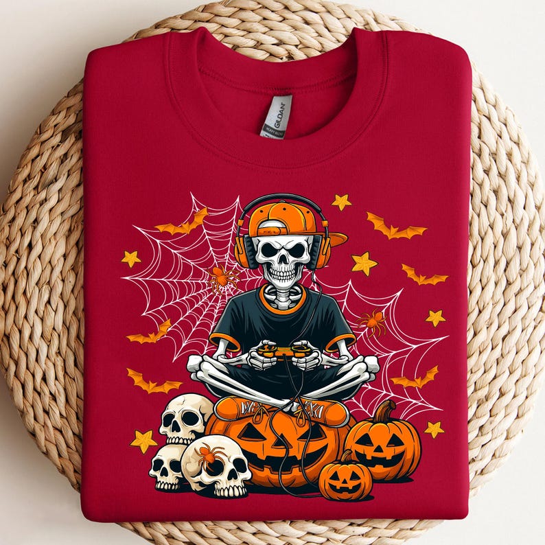 May include: A red sweatshirt with a Halloween design. The graphic depicts a skeleton wearing headphones and holding a game controller, positioned in front of pumpkins, bats, spiders, and a spiderweb. The design uses orange, black, and white.