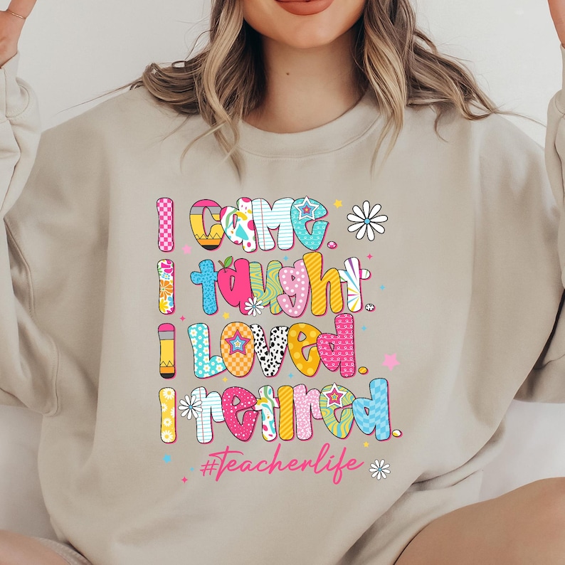 May include: Beige sweatshirt with the text "I Came, I Taught, I Loved, I Retired" in colorful, bubble letters. The design includes flowers and stars, with the hashtag #Teacherlife.