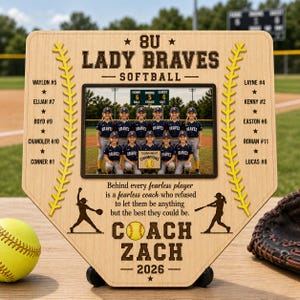 May include: A light wood home plate-shaped plaque featuring a team photo of the 8U Lady Braves softball team. The plaque includes yellow softball stitching graphics, player names with numbers, and text honoring Coach Zach for the 2026 season.