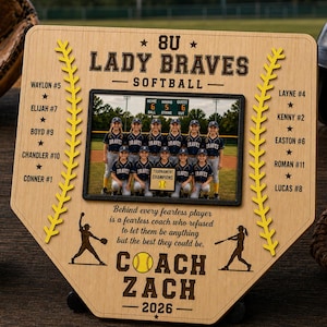 May include: A wooden baseball home plate-shaped plaque with a team photo, text, and yellow baseball stitching. The text reads "8U Lady Braves Softball" and "Coach Zach 2026". A softball and glove are visible.