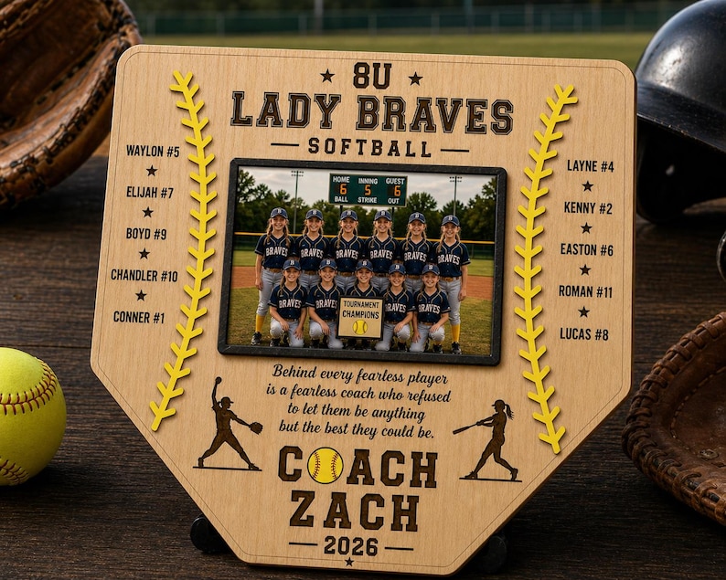 May include: A wooden baseball home plate-shaped plaque with a team photo, text, and yellow baseball stitching. The text reads "8U Lady Braves Softball" and "Coach Zach 2026". A softball and glove are visible.