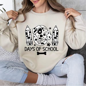 May include: A cream-colored sweatshirt with a black and white cartoon dalmatian dog wearing a graduation cap and holding a pencil. The text "100 Days of School" is printed below the dog.