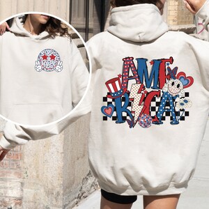 May include: A white hoodie with a red, white, and blue "AMERICA" graphic on the back. The graphic includes a smiley face, stars, a lightning bolt, a checkered pattern, and a red, white, and blue top hat. The front of the hoodie has a white and blue smiley face with stars for eyes.