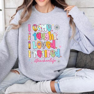 May include: A light gray sweatshirt with the text "I CAME I TAUGHT I LOVED I RETIRED" in colorful, patterned letters. The phrase "#Teacherlife" is below the text, with flower and star accents.