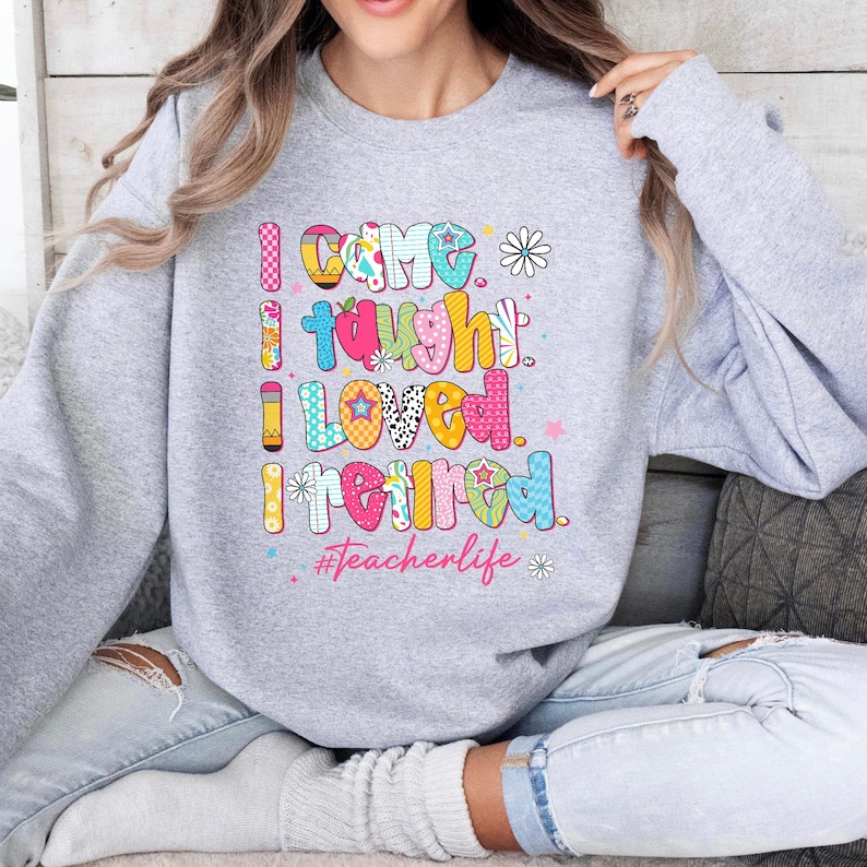 May include: A light gray sweatshirt with the text "I CAME I TAUGHT I LOVED I RETIRED" in colorful, patterned letters. The phrase "#Teacherlife" is below the text, with flower and star accents.