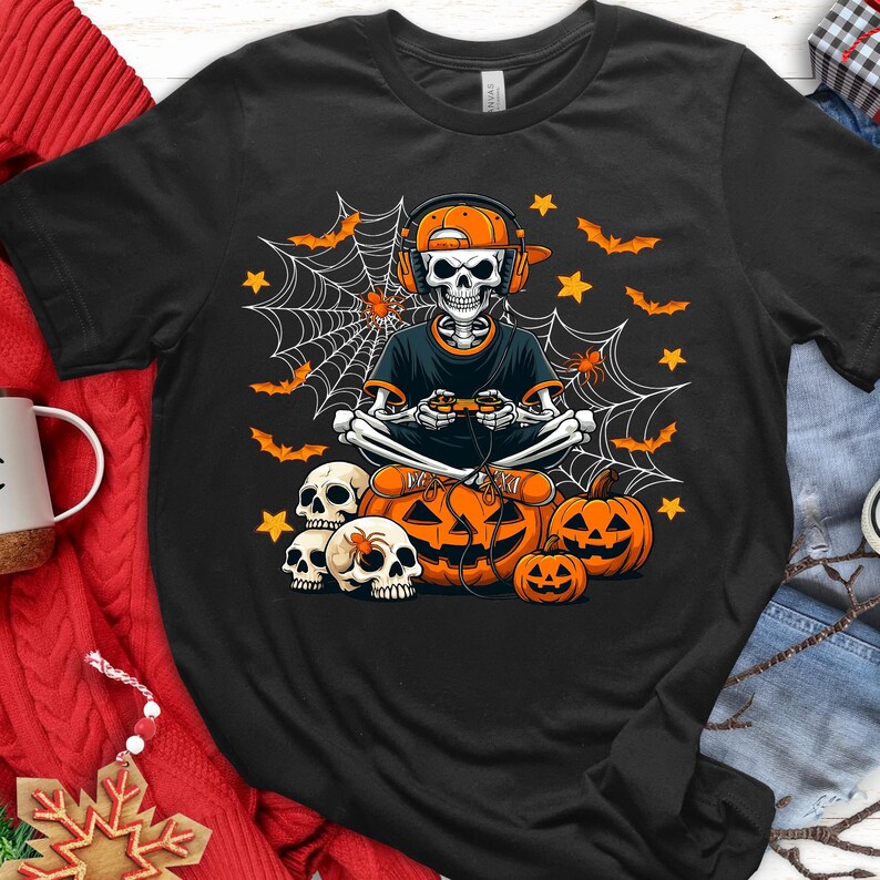 May include: Black t-shirt with a Halloween-themed design. It features a skeleton wearing a headset and cap, playing a video game. The design includes pumpkins, bats, spiderwebs, and stars.
