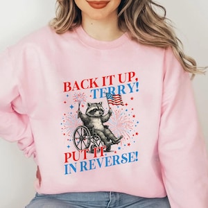 May include: Pink sweatshirt with the text "BACK IT UP, TERRY! PUT IT IN REVERSE!" featuring a raccoon in a wheelchair holding a sparkler and an American flag. The design includes fireworks and stars.