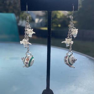 May include: Silver-toned dangle earrings featuring a chain design with small white bone-shaped charms and iridescent beads. Each earring has a small, white and green enameled teapot charm with floral accents.