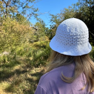 May include: A person wearing a white crochet bucket hat and a lavender t-shirt, standing in a grassy field with trees in the background.