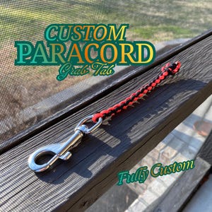Puede incluir: Una agarradera de paracord personalizada en rojo y negro, con un cierre metálico. El texto "CUSTOM PARACORD Grab Tab" y "Fully Custom" son visibles. El artículo se muestra sobre una superficie de madera oscura.