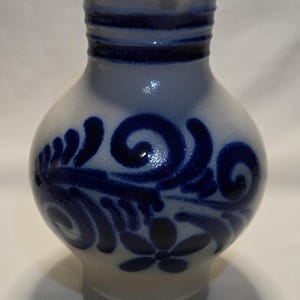 May include: A stoneware pitcher with a light gray base and body, featuring dark blue floral and swirl designs. The neck has horizontal blue bands, and a spout is visible at the top. The pitcher has a bulbous shape.