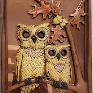 May include: A framed wall decoration featuring two yellow owls perched on a branch with brown leaves. The owls have large black eyes and are surrounded by a brown wooden frame.