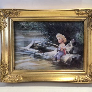 May include: A framed painting depicts a child fishing from a log in a river. The child wears a straw hat and overalls. The ornate gold frame adds a classic touch to the artwork.