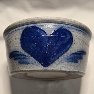 May include: A small, round, stoneware bowl with a blue heart design and wing accents. The bowl is primarily gray with a dark blue heart in the center. The heart has a textured appearance, and the wings are painted in a lighter shade of blue.