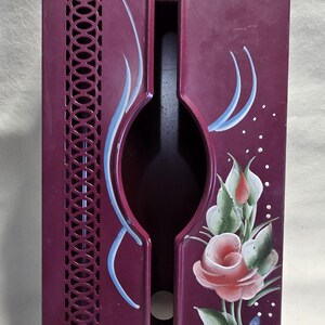 Pode incluir: Um porta-garrafas de vinho de metal bordô com um design floral decorativo. O suporte apresenta uma forma oval recortada e um design de fenda vertical no lado esquerdo. Rosas pintadas à mão e detalhes azuis adornam a frente.