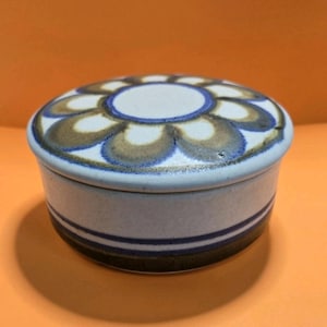 May include: A ceramic trinket box with a floral design on the lid. The lid features a white center surrounded by blue and brown petal shapes. The box has a white base with blue and brown stripes.