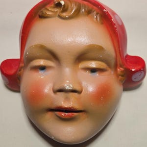 Puede incluir: Adorno de pared de cerámica vintage con forma de rostro infantil. El rostro tiene mejillas rosadas, labios rojos y ojos azules. El niño lleva un gorro rojo con lunares blancos. La cara está pintada con un acabado mate.