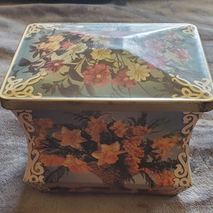 May include: A vintage tin box with a floral design. The lid features a bouquet of colorful flowers on a blue background with gold accents. The bottom of the box features a bouquet of yellow flowers on a blue background with gold accents.