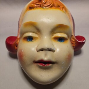 Könnte beinhalten: Ein Vintage-Puppengesicht mit blauen Augen, roten Lippen und orangefarbenem Haar. Das Puppengesicht hat einen cremefarbenen Teint mit roten Wangen und roten Akzenten an den Ohren. Das Gesicht weist leichte Gebrauchsspuren auf.