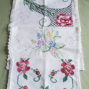 May include: Four white embroidered tea towels with different floral designs. The top towel features a peacock, the second a rose, the third a bouquet of flowers, and the bottom towel has a floral pattern with red and pink roses.
