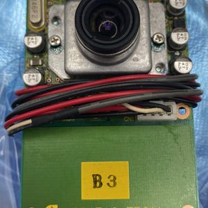 Puede incluir: Placa de circuito electrónico con una lente central, rodeada de condensadores, resistencias y otros componentes. Una sección rectangular verde en la parte inferior tiene el texto "B3" en una etiqueta amarilla y "VC047AC-EP01A" impreso debajo.