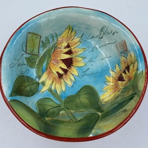 May include: A blue ceramic bowl with a red rim features a hand-painted design of sunflowers with green leaves and the text "Indian Blush Autumn" in cursive.