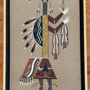Puede incluir: Una pintura de arena rectangular en un marco negro que presenta una figura estilizada con cuernos, un cuello largo y una falda estampada. La figura está adornada con plumas y diseños geométricos en tonos marrón, negro, blanco y amarillo.