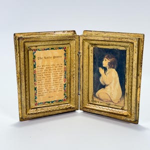 May include: A vintage, hinged, gold-framed religious diptych. One side displays a child in prayer, the other features the text of The Lord's Prayer. The frames have ornate detailing and a distressed finish, suggesting age.
