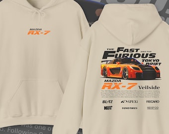 May include: A beige hoodie with a graphic print of a red and orange Mazda RX-7 sports car. The text "The Fast and the Furious Tokyo Drift" is printed above the car. The text "Mazda RX-7 Veilside" is printed below the car. The text "BLITZ NOS APEXI TOYOTIRES RECARO sparco" is printed below the car.