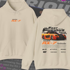 May include: A beige hoodie with a graphic print of a red and orange Mazda RX-7 sports car. The text "The Fast and the Furious Tokyo Drift" is printed above the car. The text "Mazda RX-7 Veilside" is printed below the car. The text "BLITZ NOS APEXI TOYOTIRES RECARO sparco" is printed below the car.