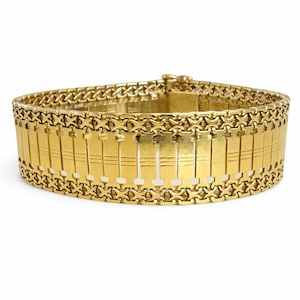May include: A gold-colored bracelet with a wide band design. The bracelet features a series of rectangular links with vertical lines, connected by intricate chain links. The clasp is visible at the top. A piece of jewelry.