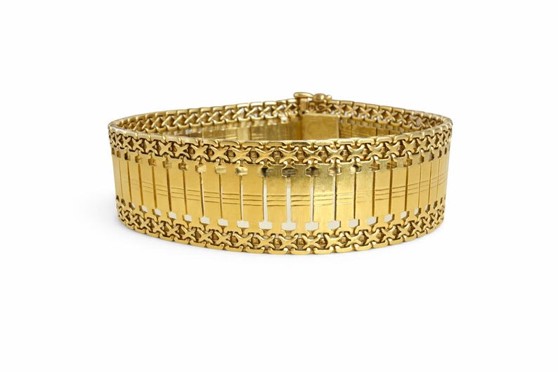 May include: A gold-colored bracelet with a wide band design. The bracelet features a series of rectangular links with vertical lines, connected by intricate chain links. The clasp is visible at the top. A piece of jewelry.