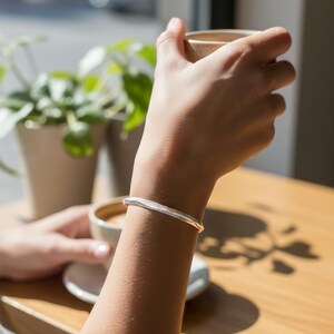 Puede incluir: Un brazalete plateado y dorado con un diseño texturizado. El brazalete se lleva en un brazo, con una taza de café en el fondo. El brazalete es un accesorio elegante.
