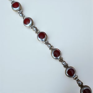 May include: A silver-toned bracelet featuring a series of round, deep red gemstones set within circular frames. The bracelet's links are composed of small, decorative beads, creating an elegant and classic design. The gemstones are evenly spaced along the length of the bracelet.