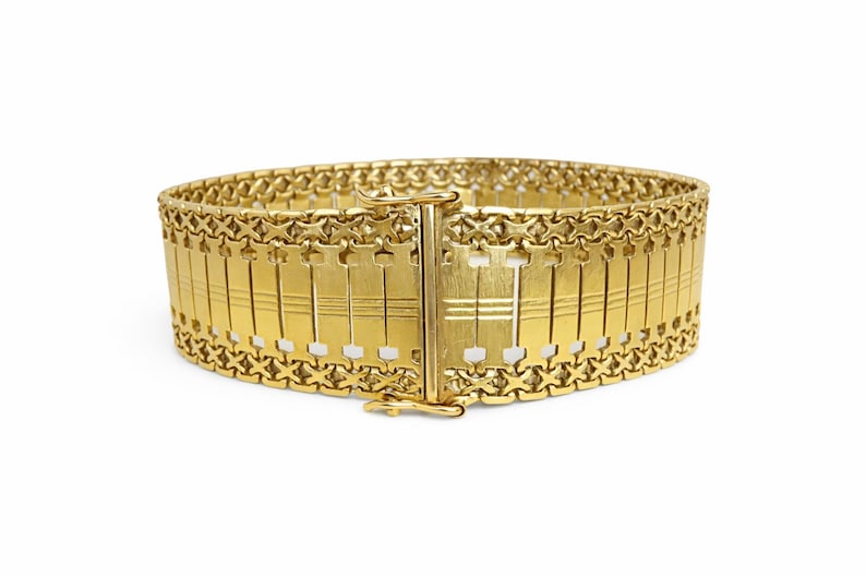 May include: A wide, gold-colored bracelet with a rectangular design. The bracelet features a series of vertical bars connected by a decorative chain. The clasp is centered and secures the bracelet. The jewelry is set against a white background.