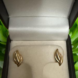 May include: A pair of gold stud earrings in a black jewellery box. Each earring features a stylized leaf design. The earrings are displayed on a cream fabric. The box is open, revealing the earrings.
