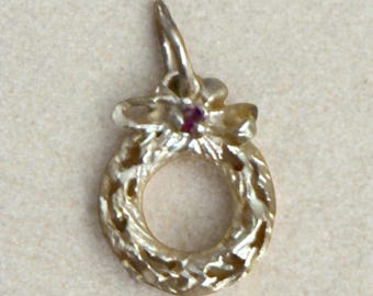 May include: A gold-toned charm in the shape of a Christmas wreath with a red jewel in the centre.