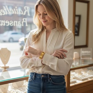 May include: A gold-colored bracelet with a textured design is worn on a wrist. The person is wearing a white blouse and blue jeans. Jewelry store display cases are visible in the background.