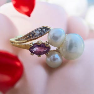 May include: A gold ring with three pearls and a purple gemstone. The ring is set on a white background.