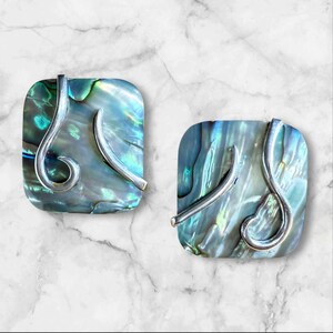 May include: A pair of square abalone shell earrings with silver metal detailing. The earrings feature a swirling design and iridescent blue, green, and grey tones. They are set against a white marble background.