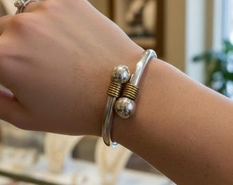 Puede incluir: Un brazalete plateado con dos detalles esféricos y detalles de alambre dorado. La pulsera se lleva en la muñeca, mostrando su diseño curvo y acabado pulido. La joya se exhibe en una joyería.