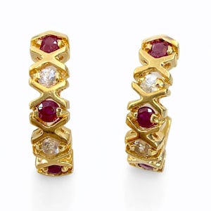 Puede incluir: Un par de pendientes de aro dorados con gemas rojas y claras alternadas. Las gemas están engastadas en un patrón geométrico repetitivo. Un accesorio elegante sobre un fondo blanco.
