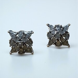May include: A pair of stud earrings with a floral design. Each earring features a cluster of small, round and pear-shaped gemstones in shades of gray and clear, set in a gold-toned metal. The earrings are displayed on a white surface.