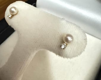 May include: A pair of pearl stud earrings with small diamond accents. The earrings feature a light cream-colored pearl and a small, clear diamond. The earrings are set on a white display.