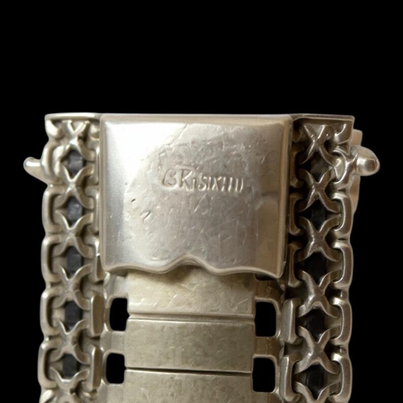May include: Close-up of a silver bracelet clasp with the engraved text "GR Sixth". The bracelet features a detailed chain design with a series of rectangular links. The background is solid black.