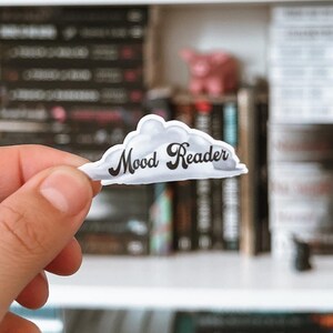 Könnte beinhalten: Ein weißer, wolkenförmiger Aufkleber mit dem Text "Mood Reader" in schwarzen Buchstaben. Der Aufkleber wird in einer Hand vor einem Bücherregal voller Bücher gehalten.
