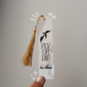 Peut inclure: Un marque-page rectangulaire transparent orné d'une silhouette de dragon noire, du texte "FLY OR DIE" et d'une icône de livre ouvert. Un pompon doré est fixé en haut par un trou, avec des gribouillages blancs d'étoiles et d'étincelles.