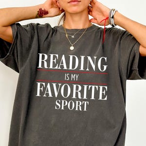 May include: Dark gray oversized t-shirt with the text "READING IS MY FAVORITE SPORT" in white. The text is separated by a red line. The shirt has short sleeves and a relaxed fit.