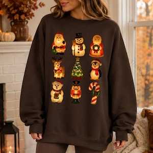 May include: A dark chocolate brown crewneck sweatshirt featuring a grid of nine Christmas-themed illustrations including Santa Claus, a snowman, a wreath-bearing figure, a reindeer, a tree, a penguin, a bear, a nutcracker, and a candy cane.